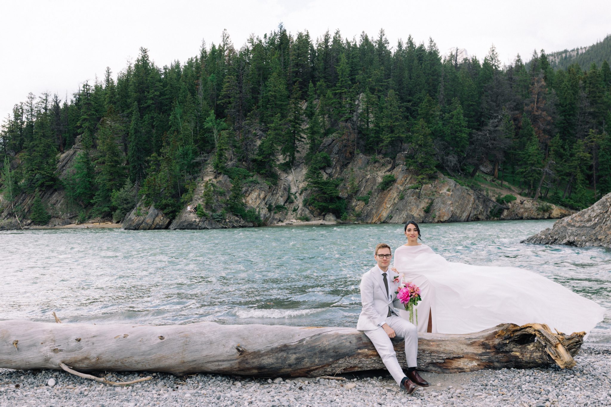 Newly married bride with a flowing cape and groom in a grey suit beside the Bow River in Banff townsite in Banff National Park in the Canadian Rockies